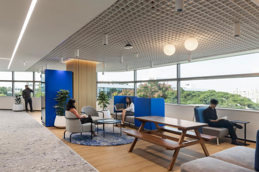 A quieter collaboration lounge: grey upholstered low chairs, deep blue acoustic divider, timber picnic bench as a long table, floor-to-ceiling glazing.