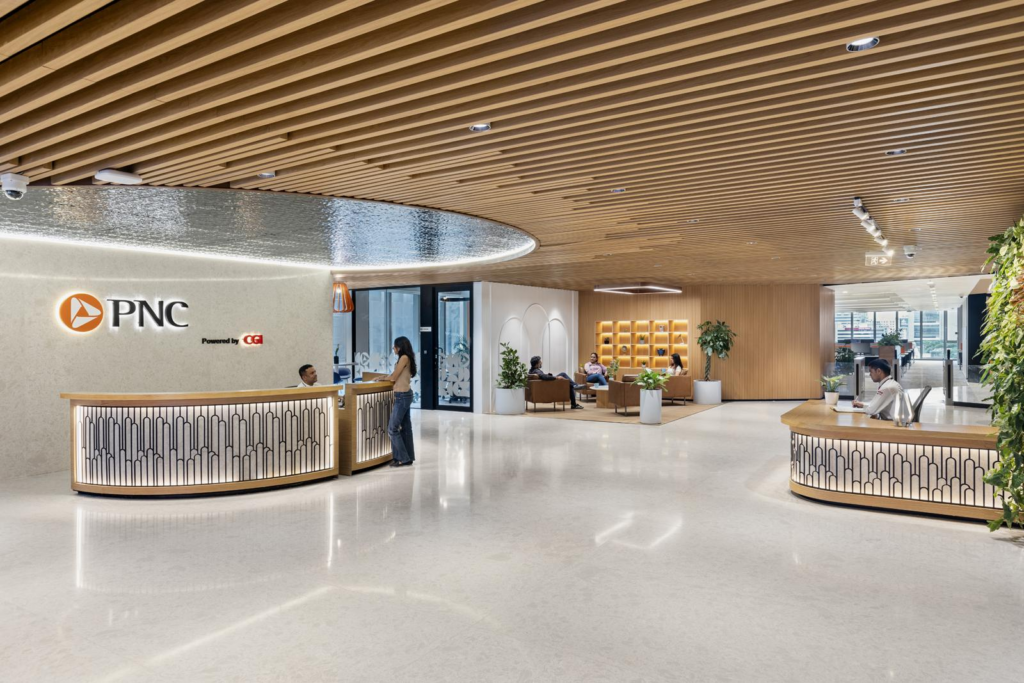 The main reception lobby: PNC brand mark and co-branded CGI signage on textured backdrop, twin curved reception desks, wood-slatted ceiling, polished terrazzo floor, planted niches.