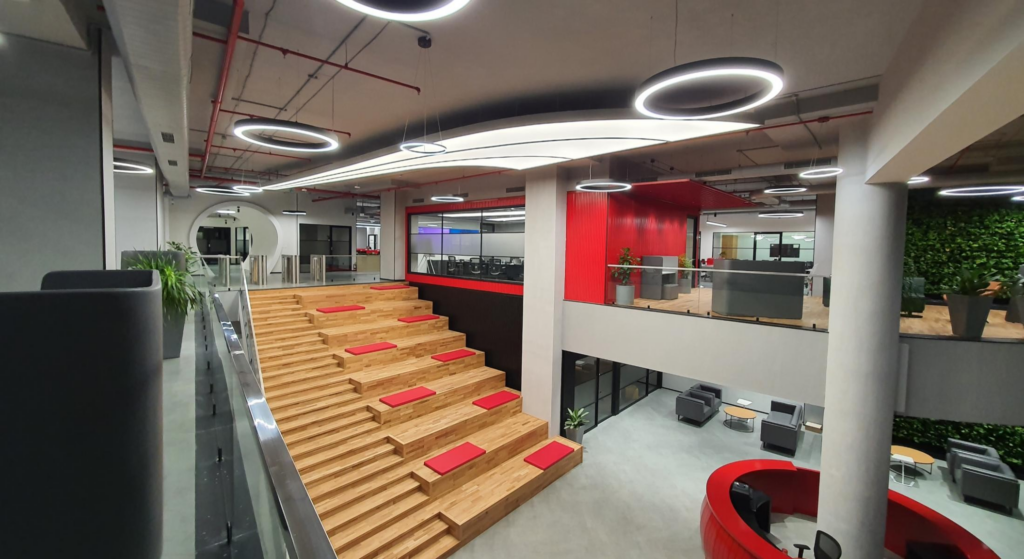 Double-height atrium: stadium timber bleachers with red cushions, circular halo light, living wall, red brand services overhead.